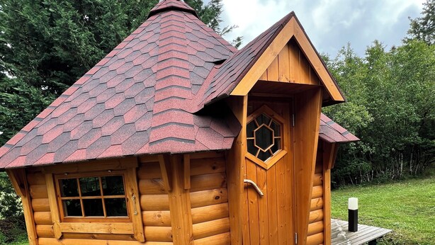 A small wooden cabin with a sloped roof, a hexagonal window, and a wooden door.