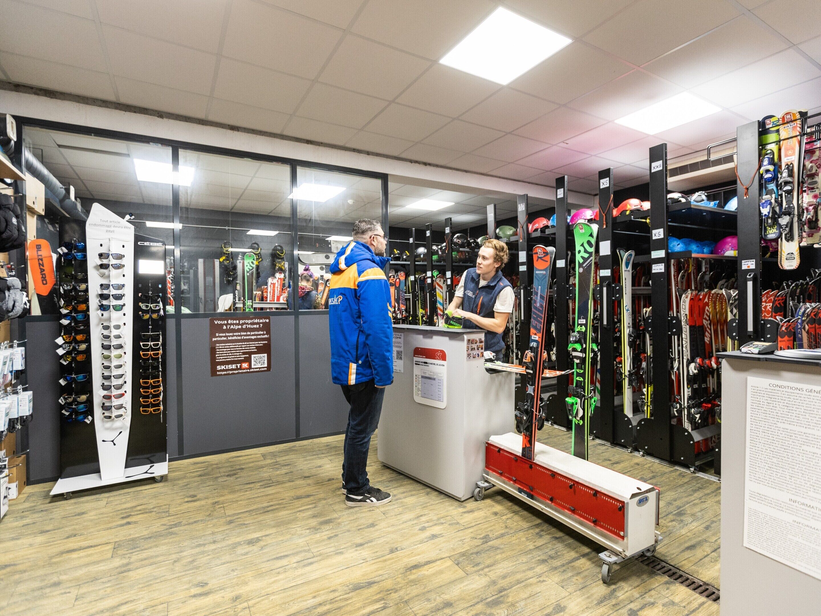 A ski equipment store with racks of skis and goggles, a counter, and a sign with information.