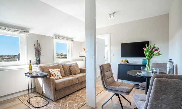 Ein modernes Wohnzimmer mit einer Couch, einem Essbereich mit Tisch und Stühlen, einem Flachbildfernseher und Blick ins Freie.