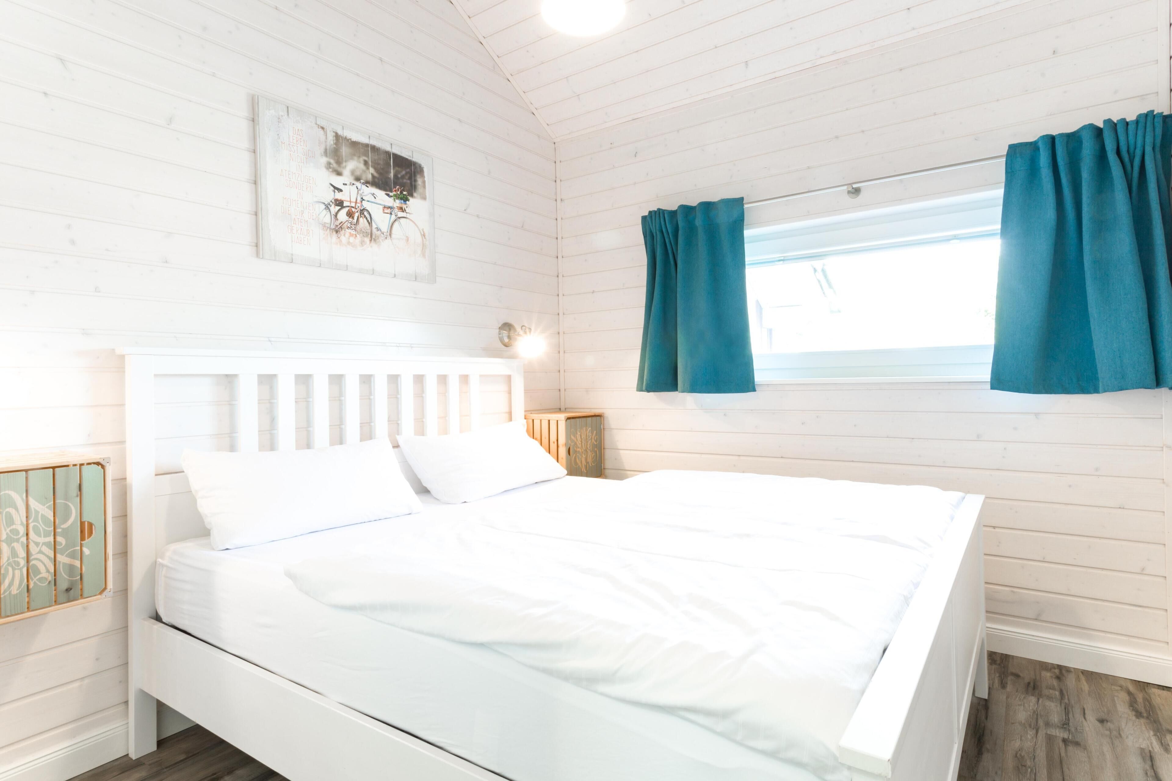 Ett vitt rum med träpanel, en säng, ett nattduksbord, en vägglampa och ett fönster med gardiner.