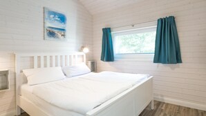 A white wooden room with a bed, a window with curtains, a wall-mounted light, and a poster. - Ferienhaus in Schönberg-Holm: Sauna, Kamin & Strandkorb inklusive (Schönberg (Holstein))