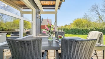 Une terrasse extérieure avec des meubles en osier, une table en verre et une vue sur une maison et un jardin.