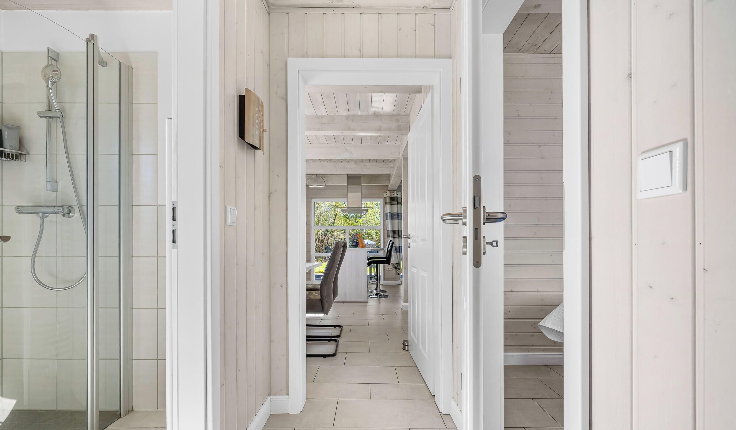 A narrow hallway with a shower, a door with a lock, and a view into a room with a chair and a window.