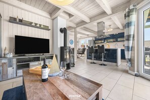Ein geräumiger Wohnbereich mit großem Flachbildfernseher, einem Holztisch mit Flasche und Gläsern, einem Kamin und einer modernen Küche im Hintergrund.