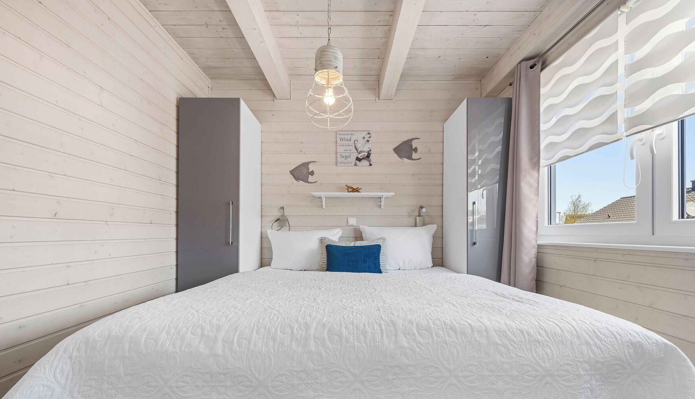 Une chambre à coucher avec un lit, un luminaire suspendu, une étagère avec des objets décoratifs et une fenêtre avec des rideaux.
