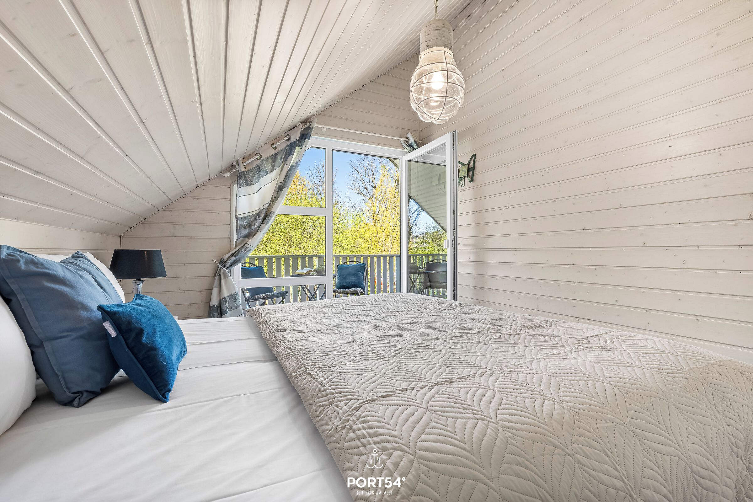 A bedroom with a bed, pillows, a hanging light fixture, and a view of the outdoors.