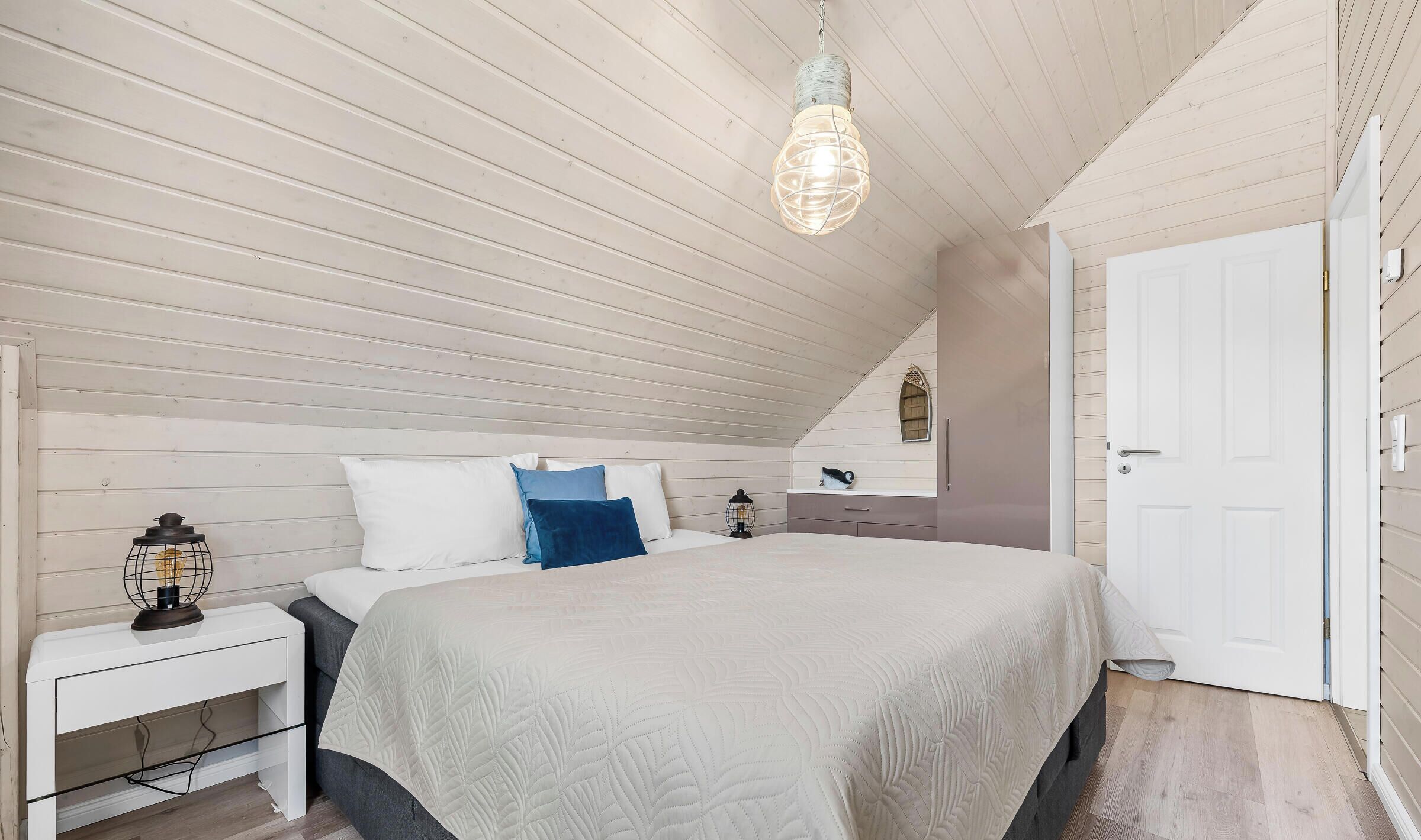 Une chambre avec un plafond en pente, un lit recouvert d’une couette blanche, une table de chevet avec une lampe et une porte.