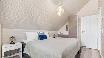 Une chambre avec un plafond en pente, un lit recouvert d’une couette blanche, une table de chevet avec une lampe et une porte.