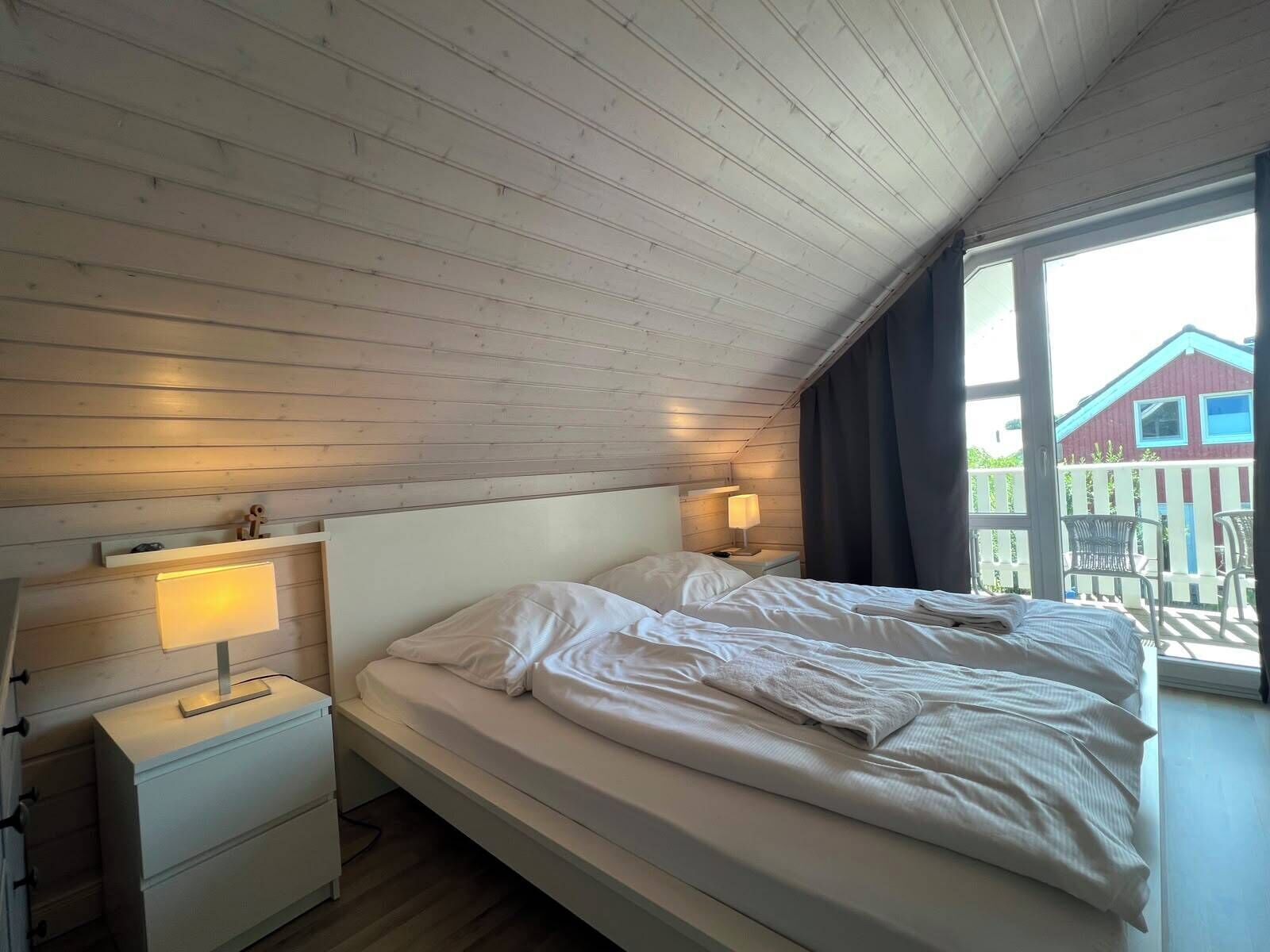 Un dormitorio con techo de madera, una cama con ropa de cama blanca, una mesita de noche con una lámpara y una ventana con vistas a un edificio rojo.