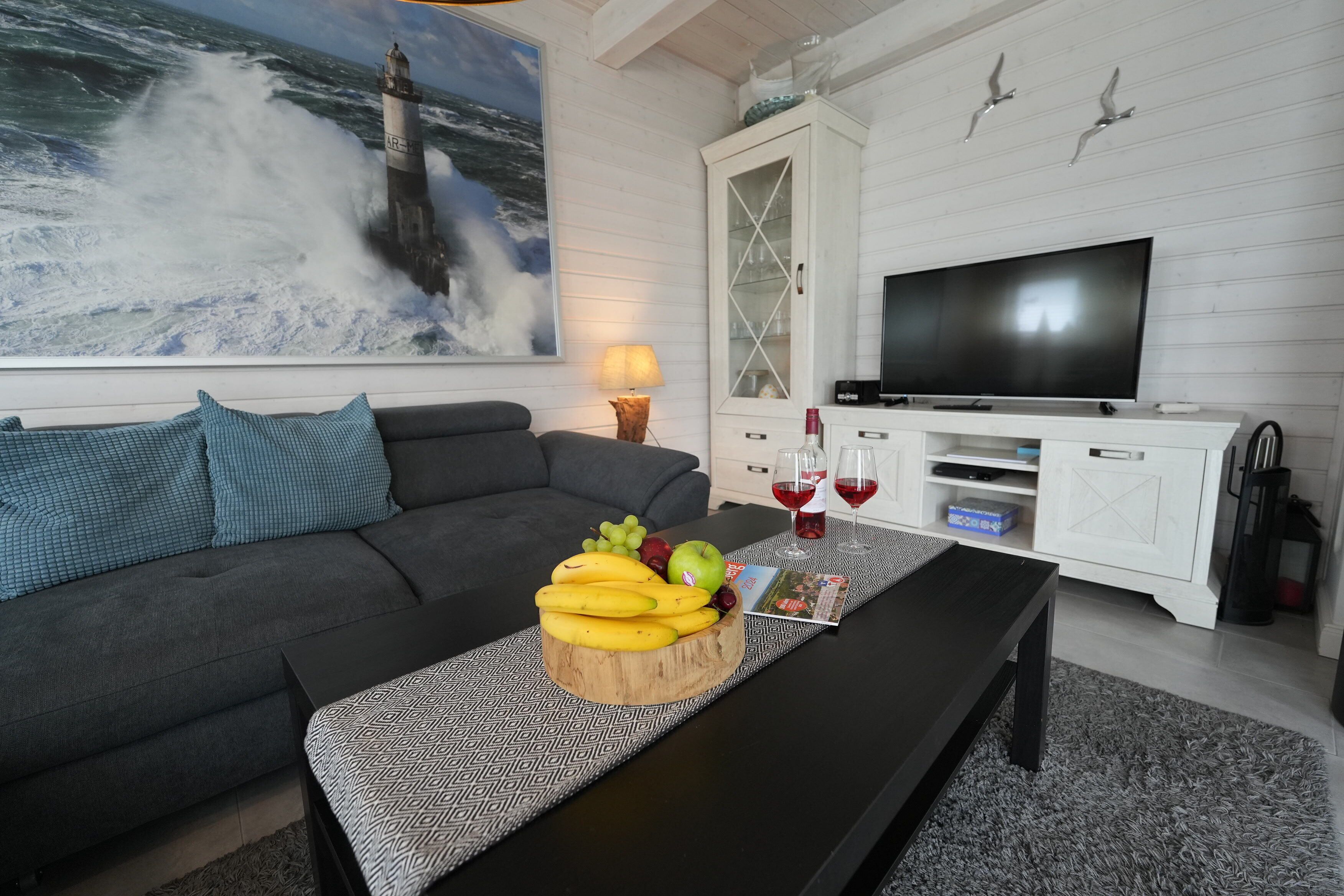 A living room with a sofa, a coffee table with fruit, a TV, and a painting of a lighthouse.
