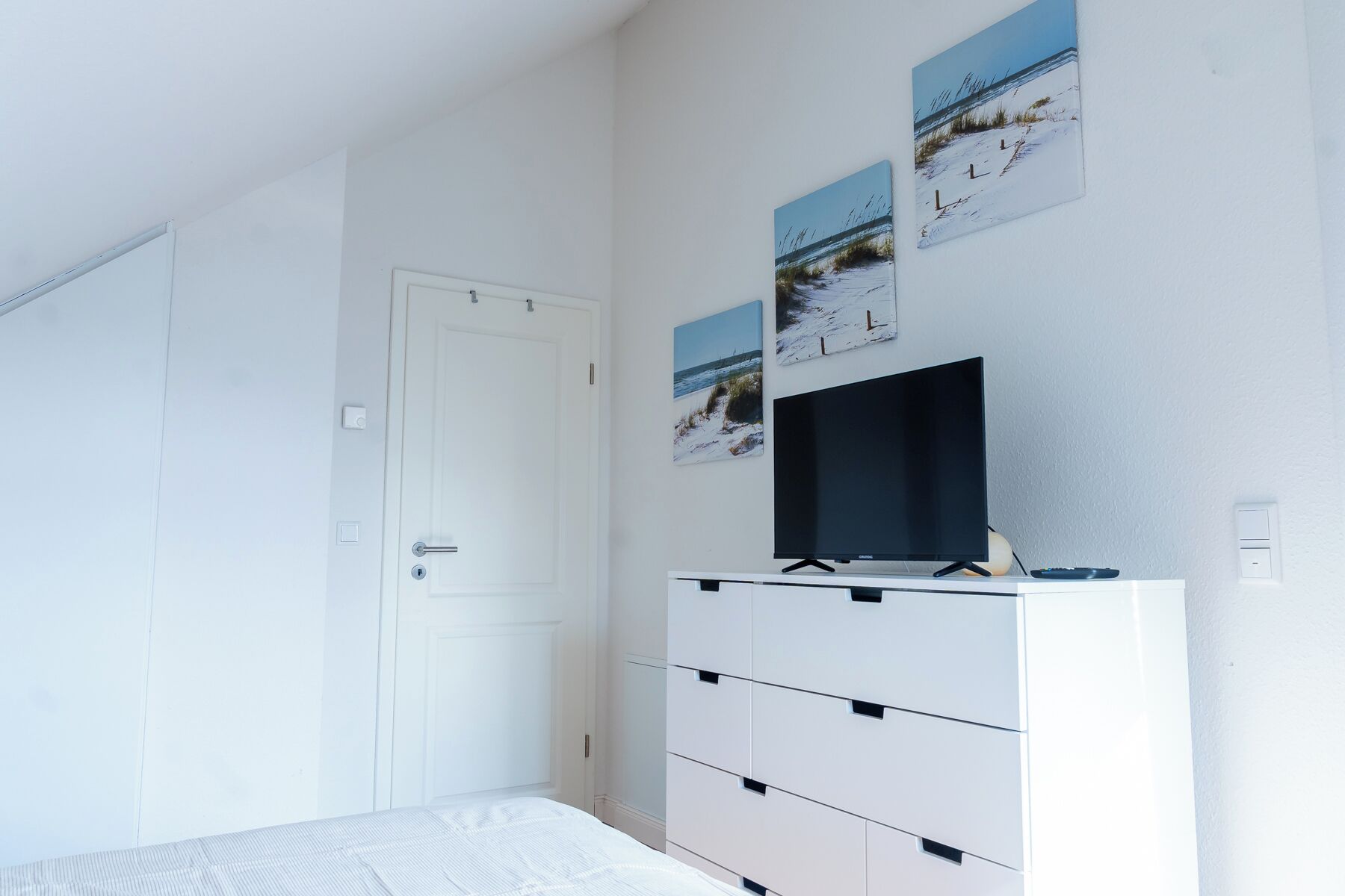A bedroom with a bed, a white dresser with drawers, a flat-screen TV, and three framed pictures on the wall.