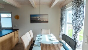 A dining area with a table set for four, surrounded by wicker chairs, a clock on the wall, and a window with patterned curtains. - Ferienhaus "Plüschmors" in Schönberg-Holm: Sauna, Kamin, 600 m Strand, Strandkor (Schönberg (Holstein))