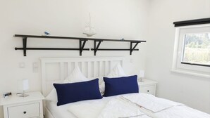 Ein Schlafzimmer mit einem weißen Bett, zwei blauen Kissen, einem weißen Kopfteil, einem Nachttisch mit einer Lampe und einem Fenster mit Blick.