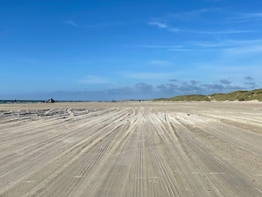 Ein weiter Sandstrand mit Radspuren, die zum Meer führen, unter einem klaren Himmel.