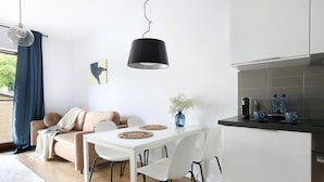 A modern kitchen with white cabinets, a dining table with chairs, a hanging black pendant light, and a window with blue curtains. - Malila Apartments City Center Pawia 34 (Kraków)