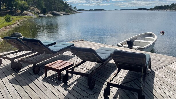 Une terrasse en bois avec des chaises longues et une petite table, donnant sur un lac paisible où un bateau est amarré à proximité.