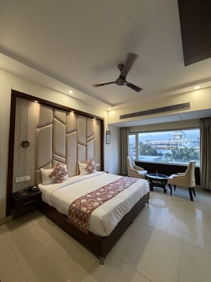 A hotel room with a large bed, a desk with a chair, a ceiling fan, and a view of the city. - Hotel Doon's Pride (Dehradun)