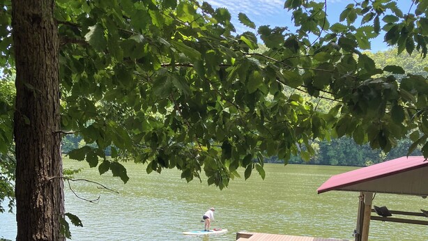 Une terrasse en bois surplombe un lac paisible, et on aperçoit au loin une personne pratiquant le paddle.