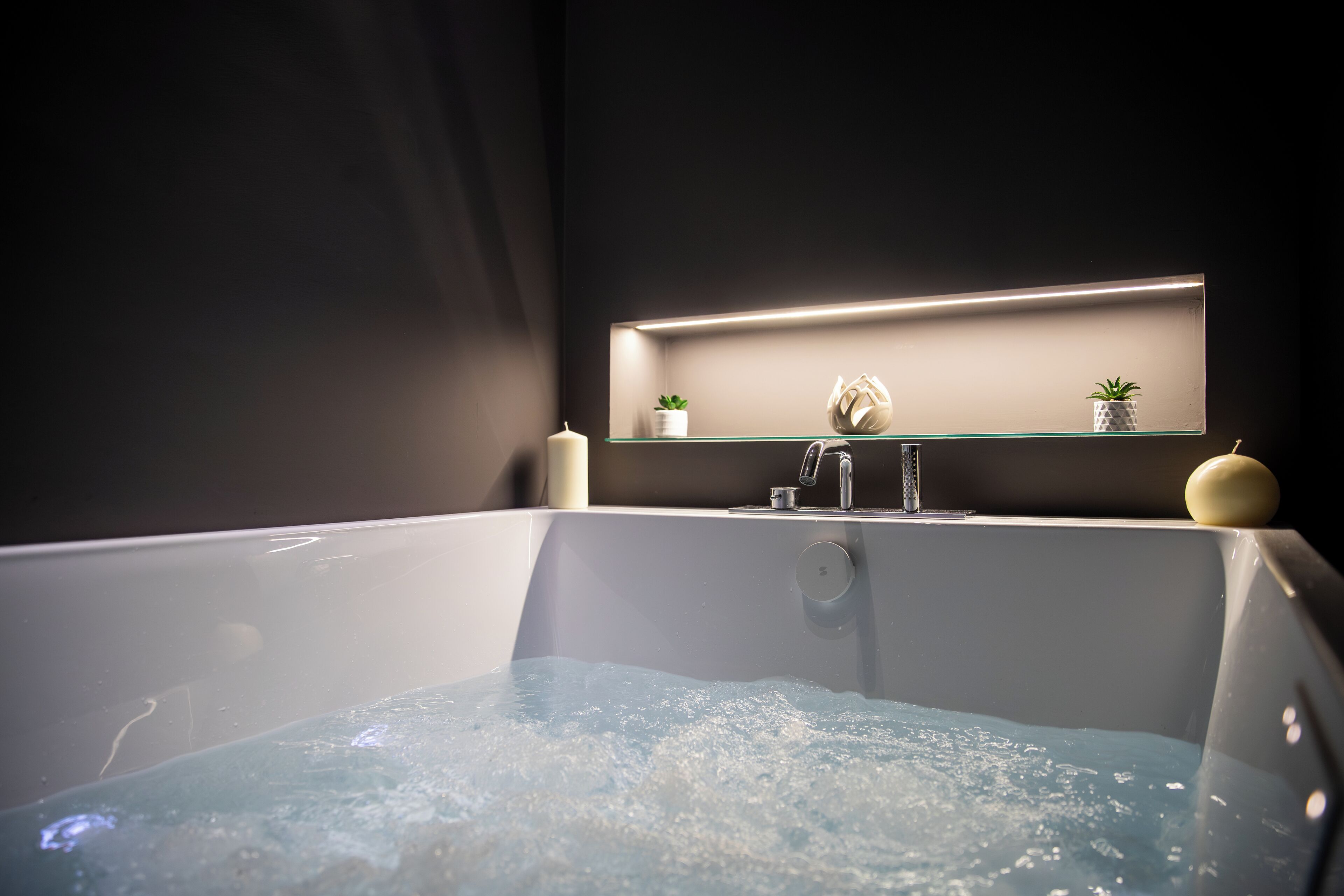 A modern bathroom with a glass-enclosed bathtub, a wall-mounted shelf with decorative items, and a visible faucet.