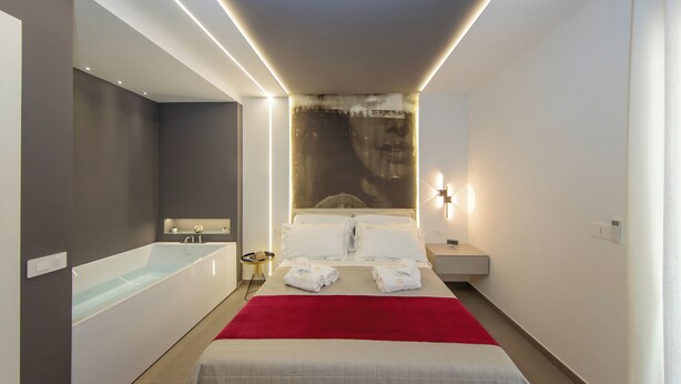 A modern hotel room with a large bed, a red throw blanket, a bathtub, and a nightstand with a lamp. - Suite 20five, Romantic Suite with In-Room Jacuzzi Hot Tub. (Bari)