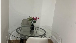Un petit coin repas avec une table en verre, une chaise blanche, une horloge au mur et un vase rempli de fleurs.