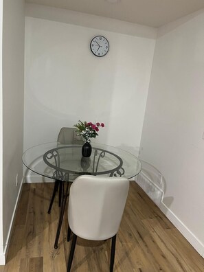 A small dining area with a glass table, a white chair, a clock on the wall, and a vase with flowers.