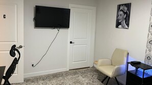 Une pièce dotée d’un téléviseur à écran plat fixé au mur, d’un fauteuil, d’un bureau avec un ordinateur et d’un tapis à motifs.