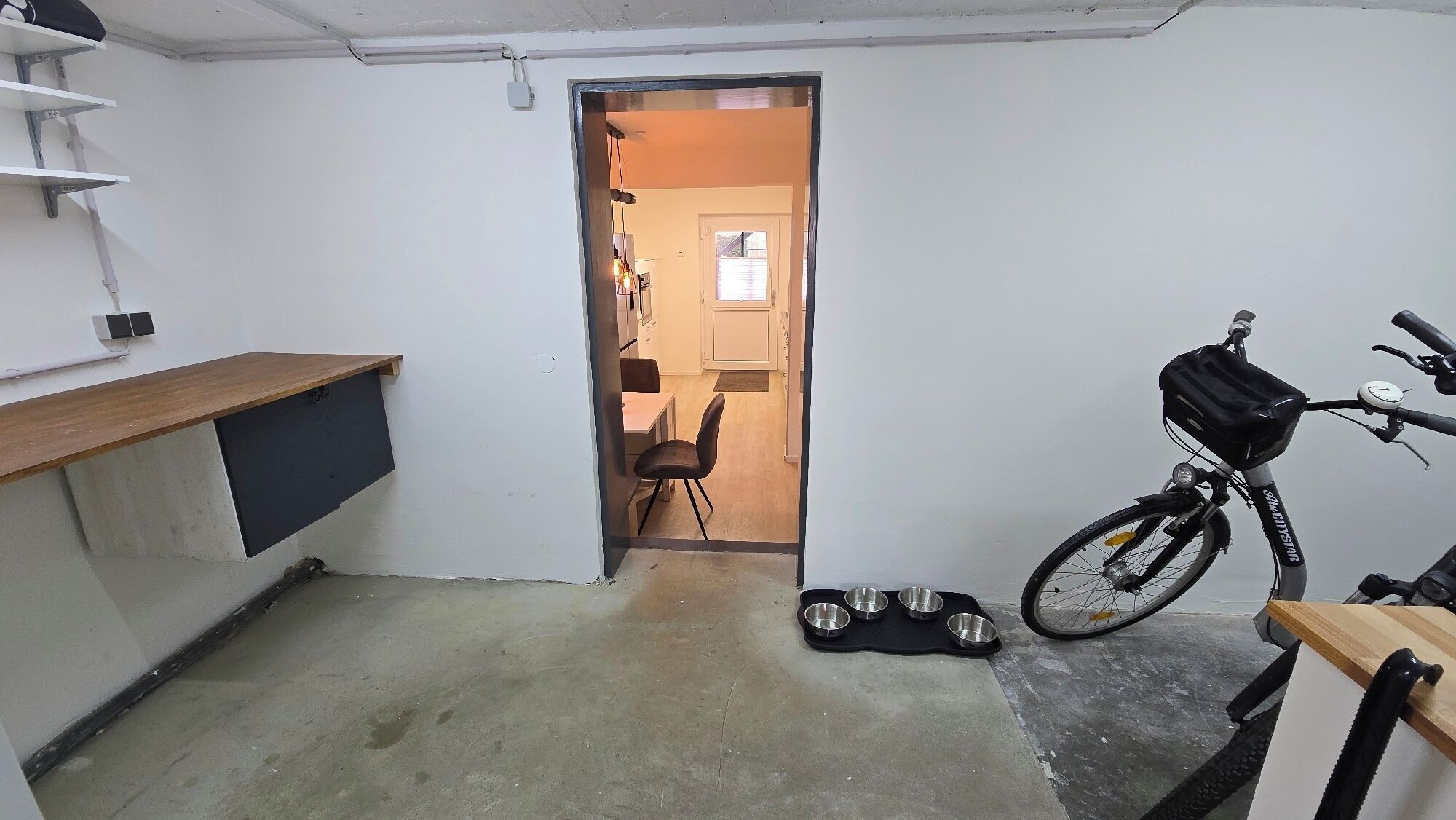 A narrow hallway with a wooden desk, a bicycle with a basket, and a door leading to a room with a chair and a table.