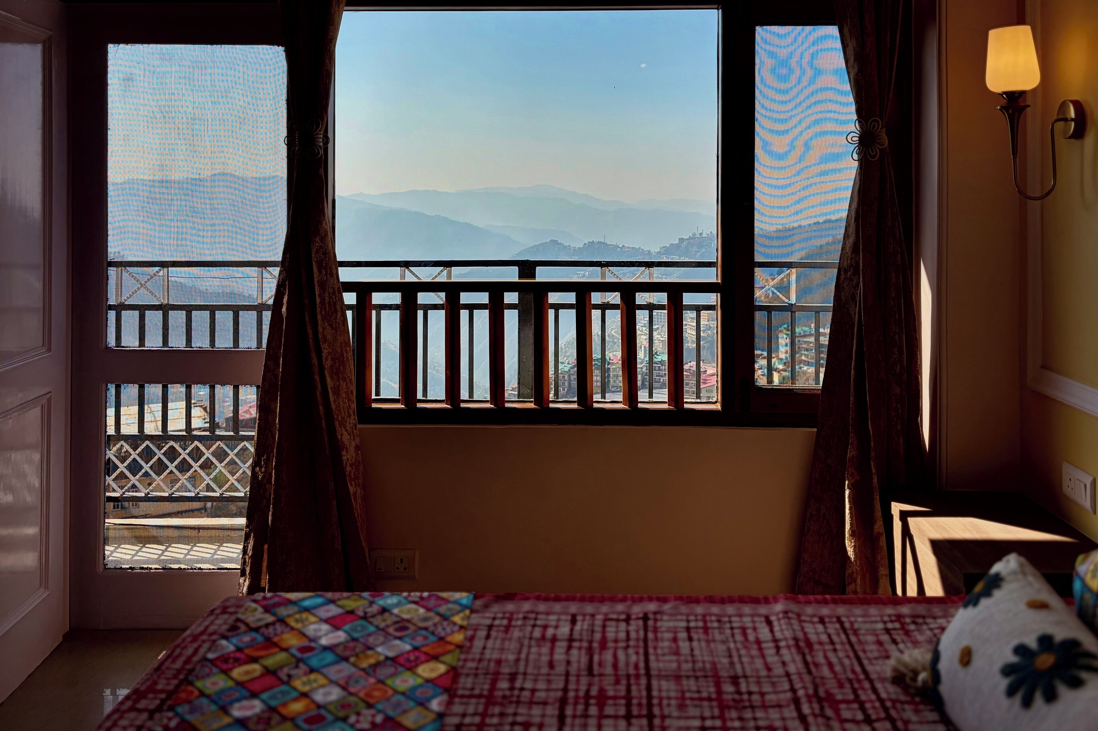 Een hotelkamer met een bed, een raam met uitzicht op bergen, een balkon met een leuning en een wandlamp.