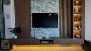 A modern living room with a flat-screen TV mounted on a marble wall, wooden paneling, a potted plant, and a shelf with decorative items. - Irra Homes • 2BHK Apartment • Panoramic Valley View (Shimla)