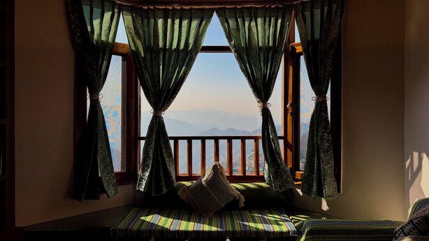 A room with a bay window, a cushioned bench, and a view of the mountains. - Irra Homes • 2BHK Apartment • Panoramic Valley View (Shimla)