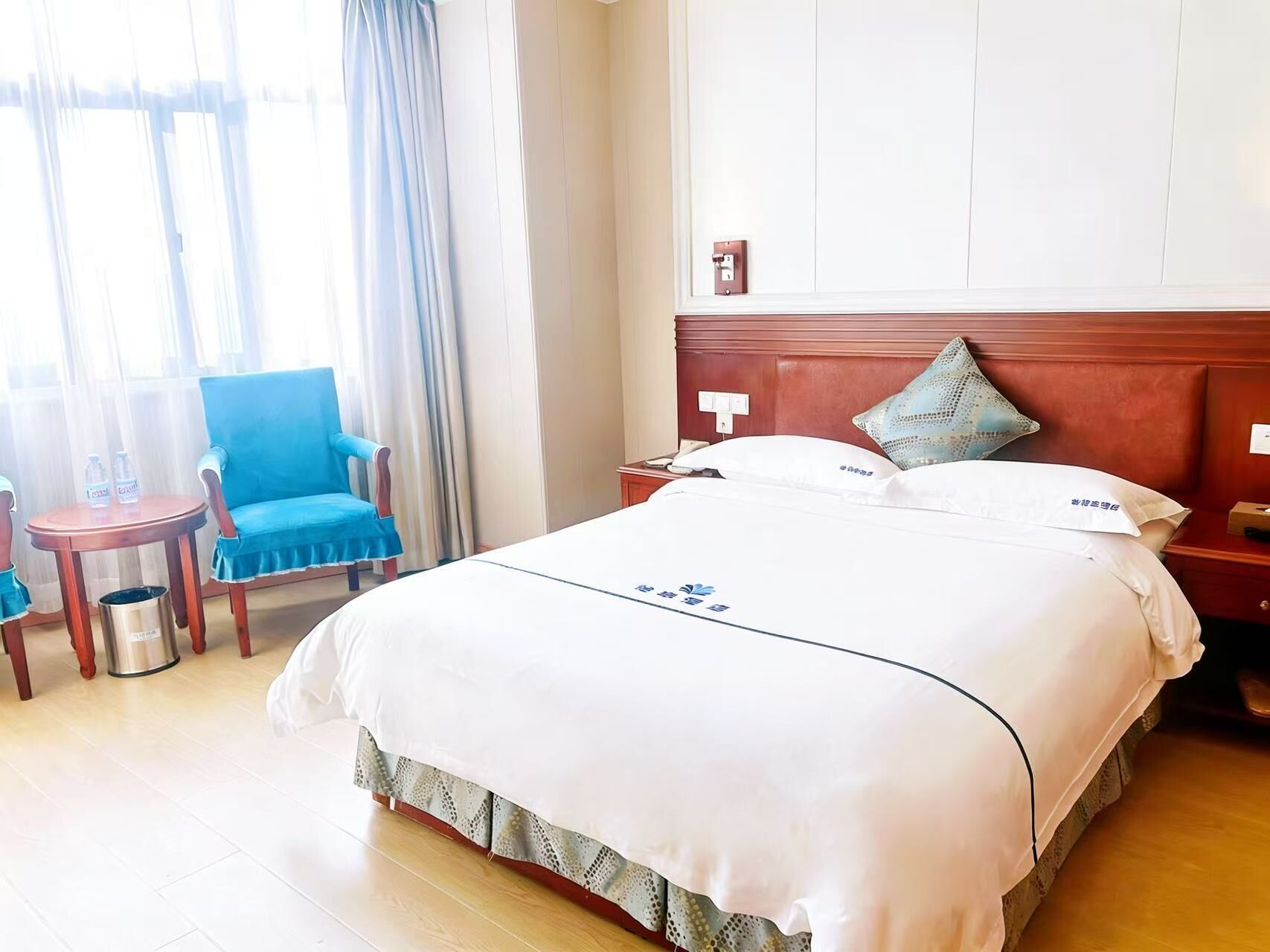 Ein Hotelzimmer mit einem großen Bett, einem Holzkopfteil, einem Nachttisch, einem Sessel und einem kleinen Tisch mit Wasserflaschen.