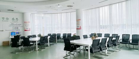 A conference room with multiple tables and chairs, large windows with blinds, and a white ceiling with recessed lighting.