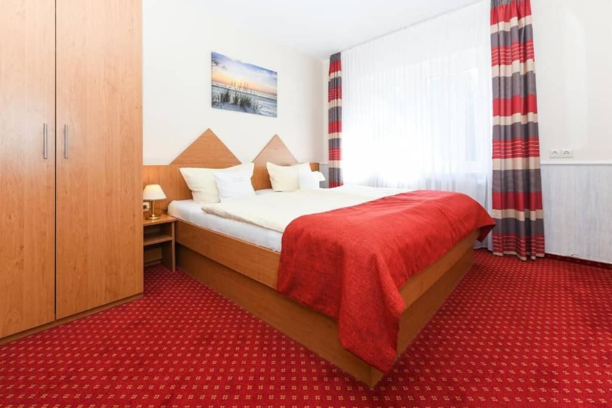 A hotel room with a wooden bed, a nightstand, a lamp, a red carpet, and a window with curtains.