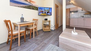A room with a dining table, chairs, a TV, a small shelf, and a kitchen area.