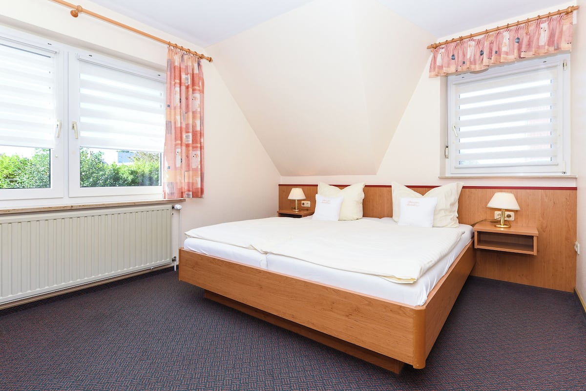 A bedroom with a large bed, two bedside tables with lamps, a window with blinds, and a radiator.