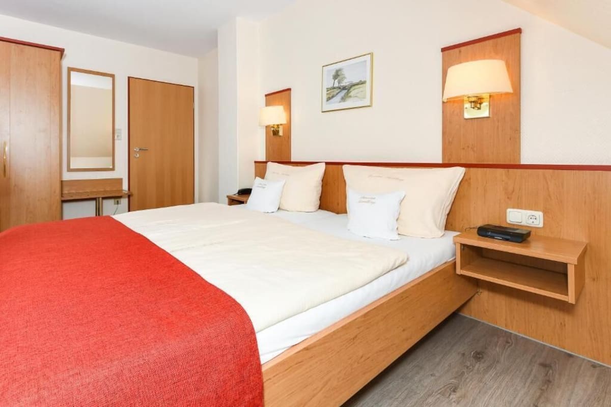 A hotel room with a bed, a bedside table, a lamp, a mirror, and a wall-mounted shelf.