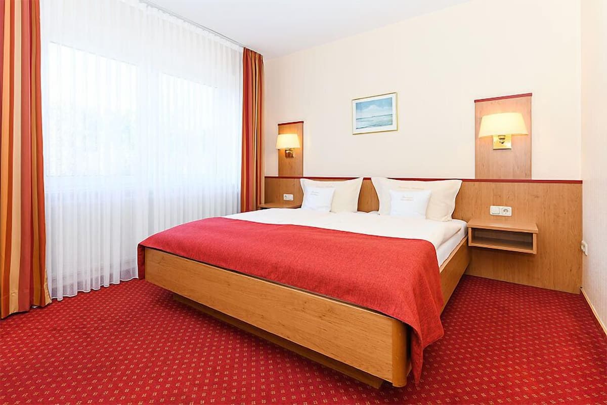 A hotel room with a bed, curtains, a wall-mounted lamp, a small table, and a painting.