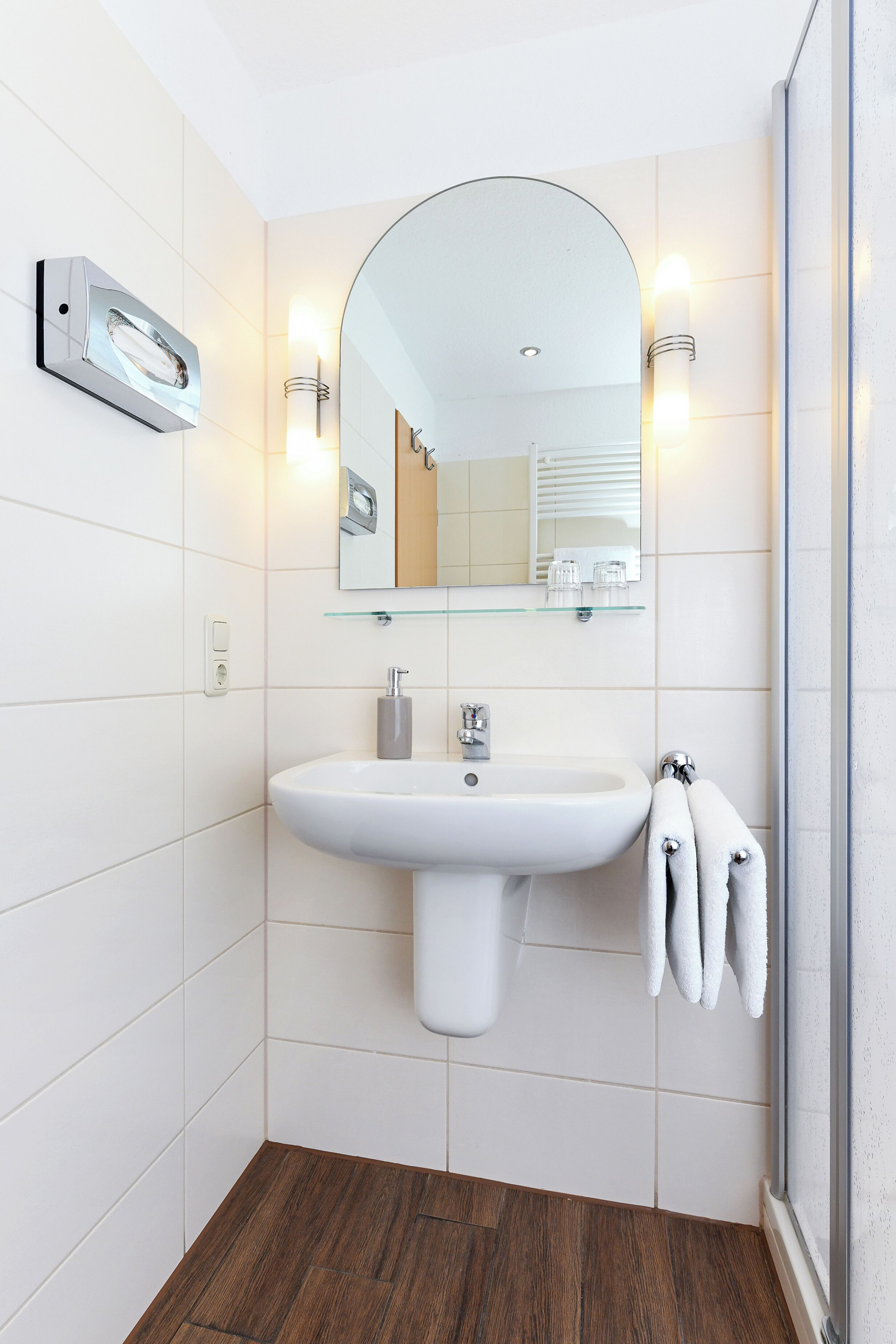 Een moderne badkamer met een wit wastafelmeubel, een glazen plank erboven, een spiegel met verlichting, en een handdoekrek.