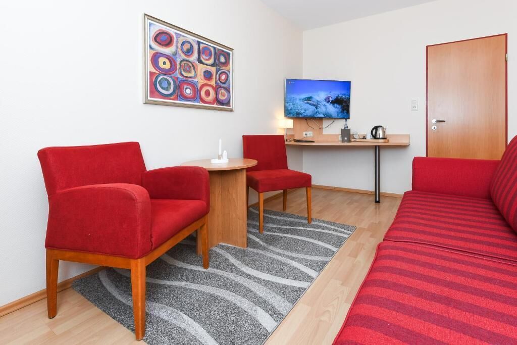 Een kamer met een rode bank, twee rode fauteuils, een klein houten tafeltje, een televisie op een meubel en een deur.