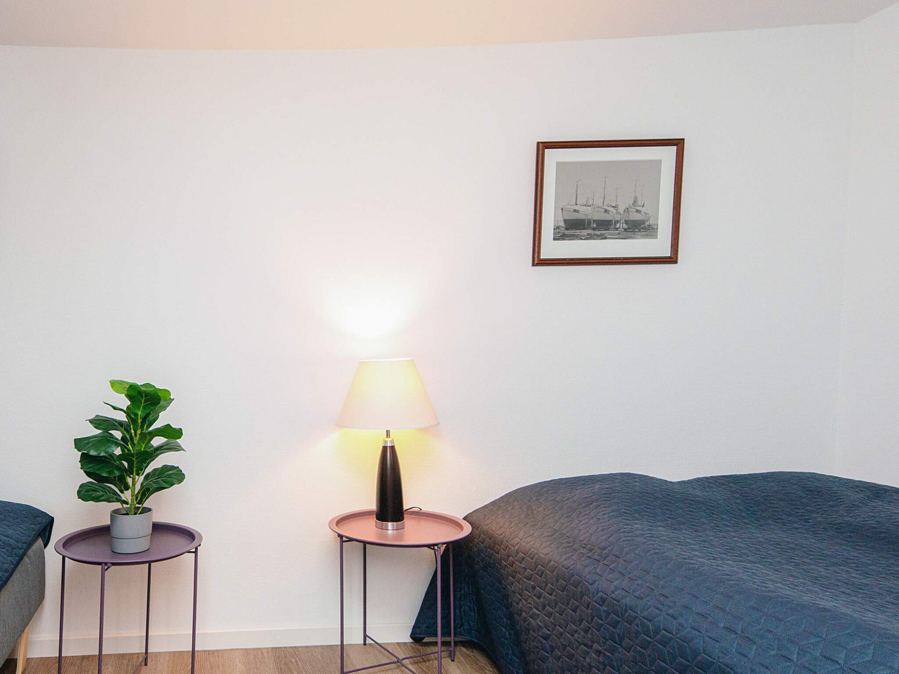 A room with a bed, a bedside table with a lamp, a framed picture on the wall, and a potted plant.