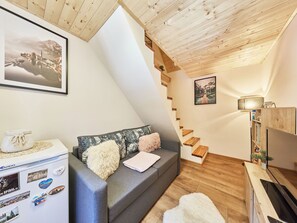 A cozy loft bedroom with a wooden ceiling, a gray sofa with decorative pillows, a small table with a white vase, a wooden TV stand, and a staircase.