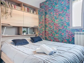 A bedroom with a bed, pillows, a bedside table with a lamp, a shelf with books and plants, and a floral wallpaper. - Dominica by Interhome (Le Barcarès)