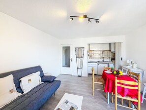 Ein modernes Wohnzimmer mit einer Couch, einem Esstisch und Stühlen, sowie einer Küche im Hintergrund und einer Deckenleuchte.