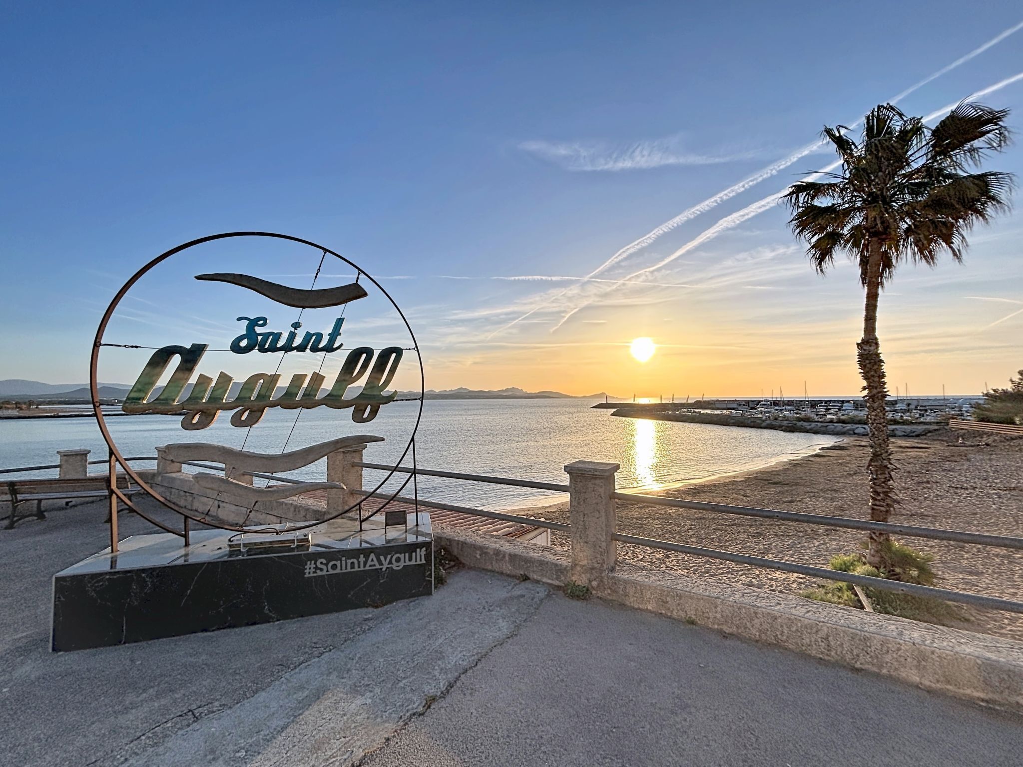 Ein Schild mit 'Saint Aygüll' und '#SaintAygüll' auf schwarzem Grund, mit Blick auf einen Strand, eine Palme und einen Sonnenuntergang.