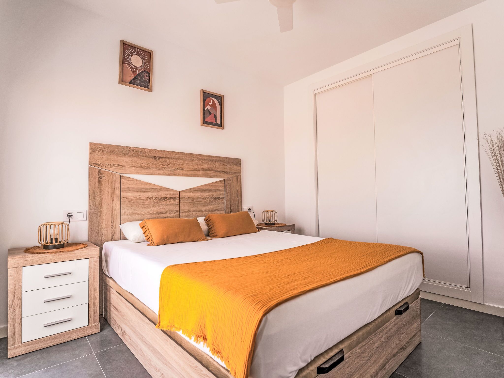 Ein Schlafzimmer mit Holz-Kopfteil, einem Bett mit weißer Bettwäsche und orangenen Kissen, einem Nachttisch mit Lampe und einem Kleiderschrank.
