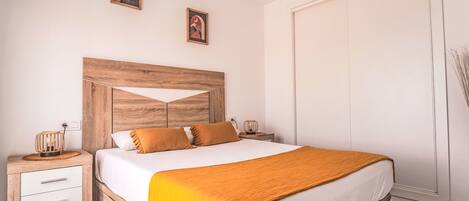 Ein Schlafzimmer mit Holz-Kopfteil, einem Bett mit weißer Bettwäsche und orangenen Kissen, einem Nachttisch mit Lampe und einem Kleiderschrank.