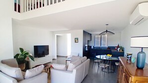 Un salon moderne comprenant un ensemble de canapés, un téléviseur, une table à manger avec des chaises, et un coin cuisine à l’arrière-plan.