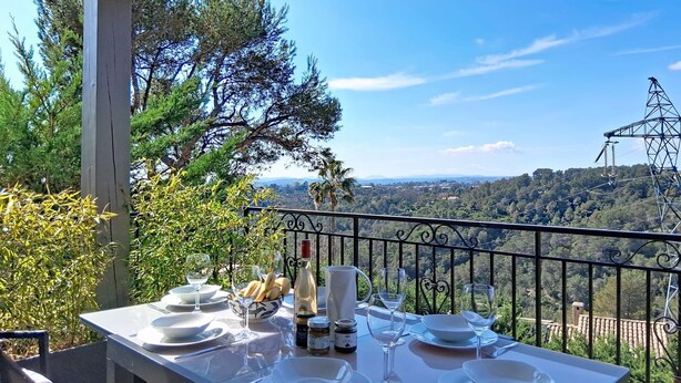 Une table de jardin, dressée avec des assiettes, des verres et des condiments, offre une vue panoramique sur un paysage verdoyant, avec des arbres et une ligne électrique éloignée.