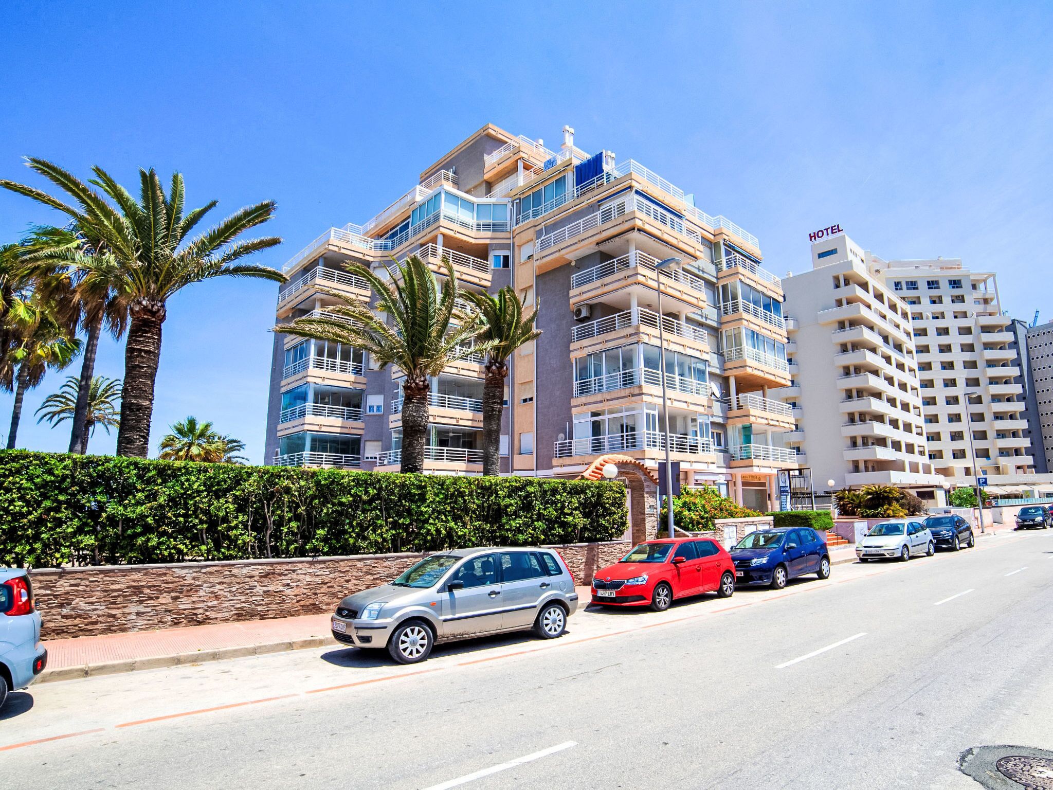 A multi-story hotel with balconies, a parking area, and palm trees.